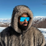 Person wearing a fur-lined hooded coat and blue sunglasses with a snowy mountain landscape in the background