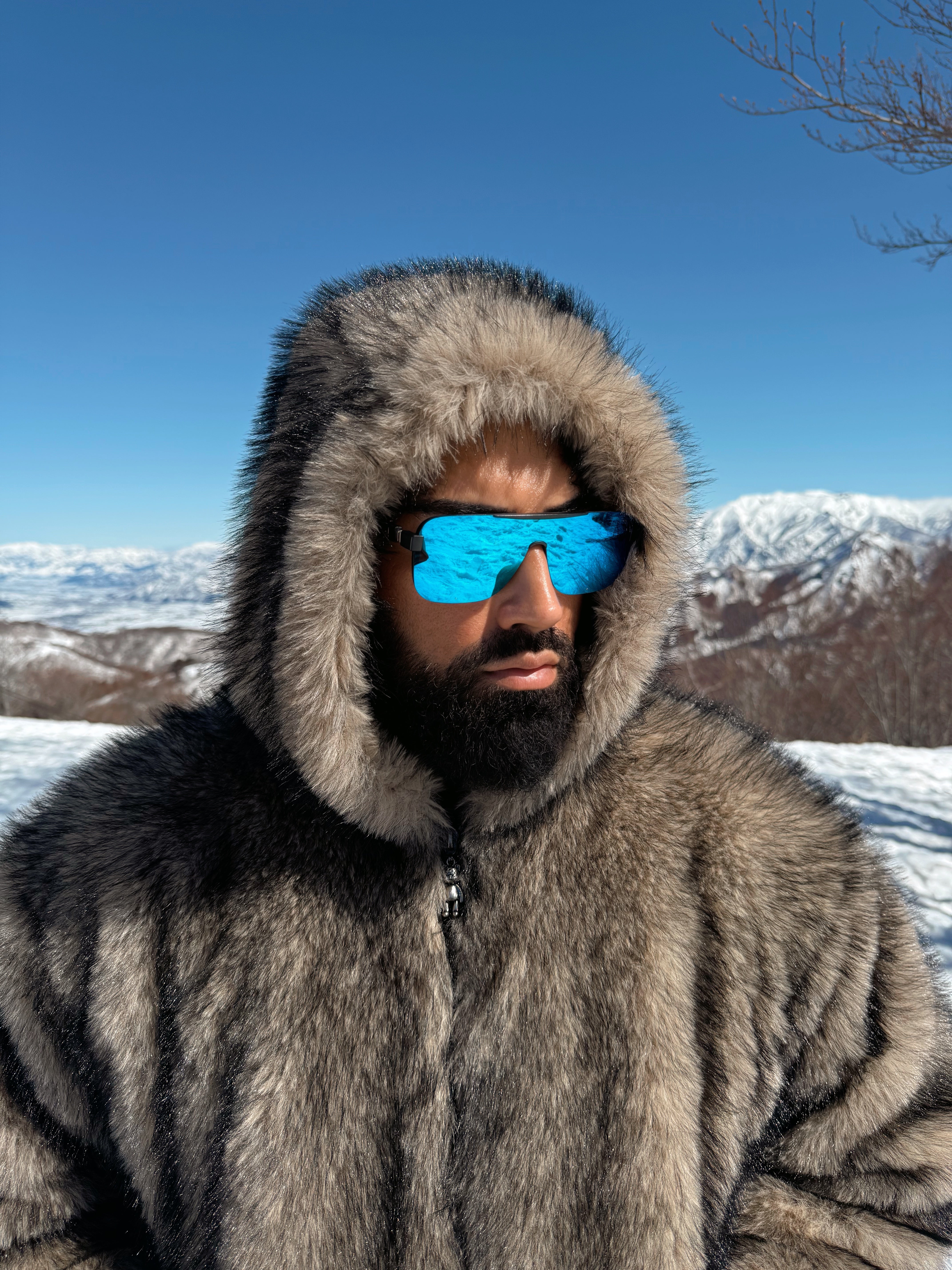 Person wearing a fur-lined hooded coat and blue sunglasses with a snowy mountain landscape in the background