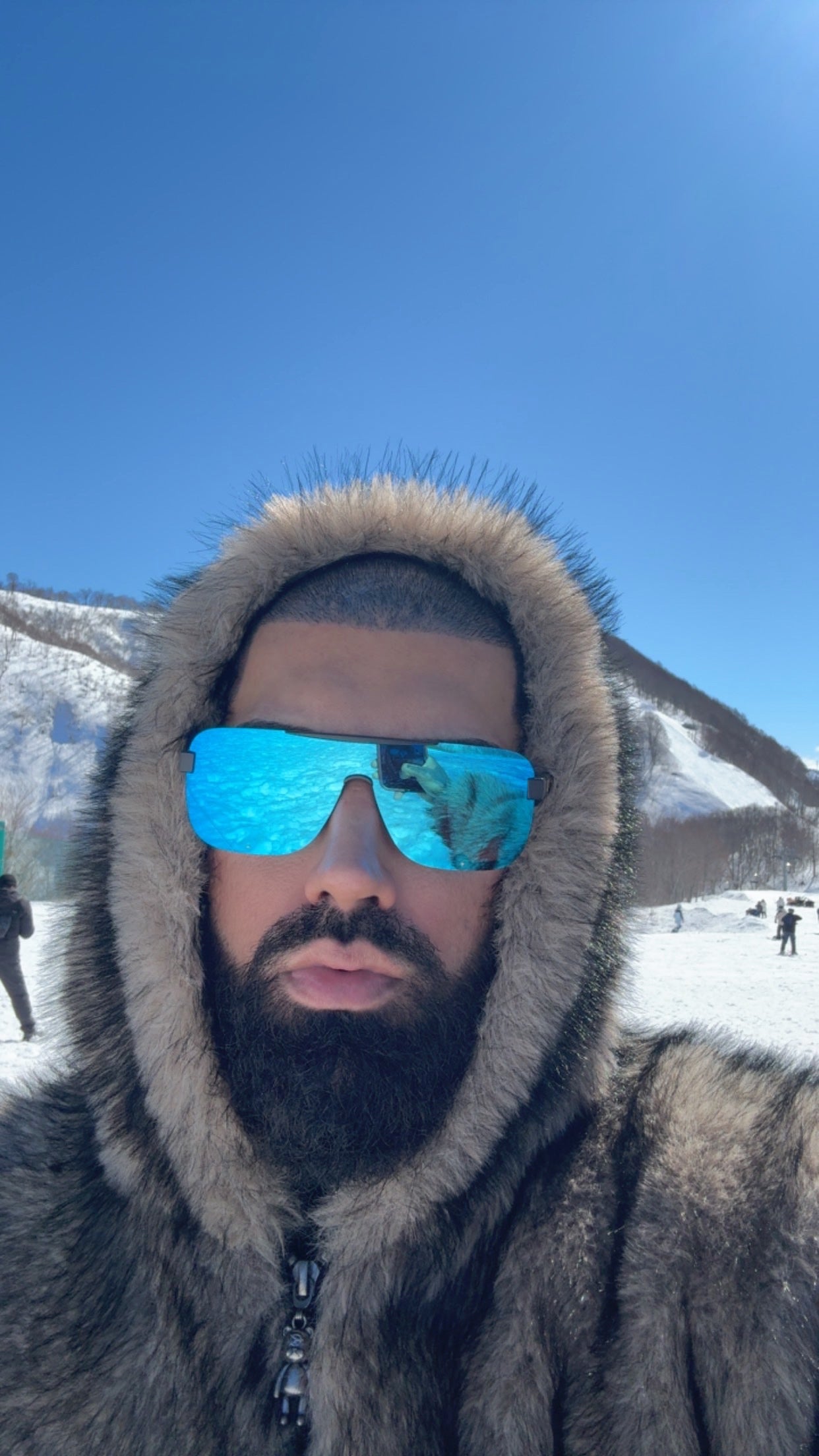 Person wearing a fur-lined hood and reflective sunglasses with mountains in the background