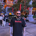 Man wearing a black 'Badfella' shirt in an outdoor setting with traditional architecture and decorations.