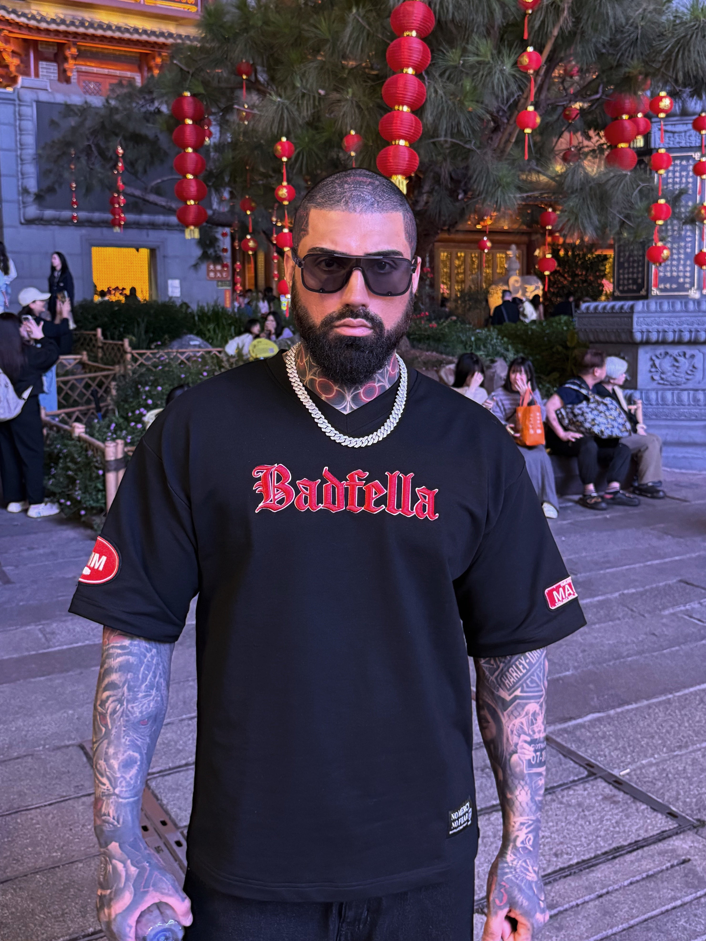 Man wearing a black 'Badfella' shirt in an outdoor setting with traditional architecture and decorations.