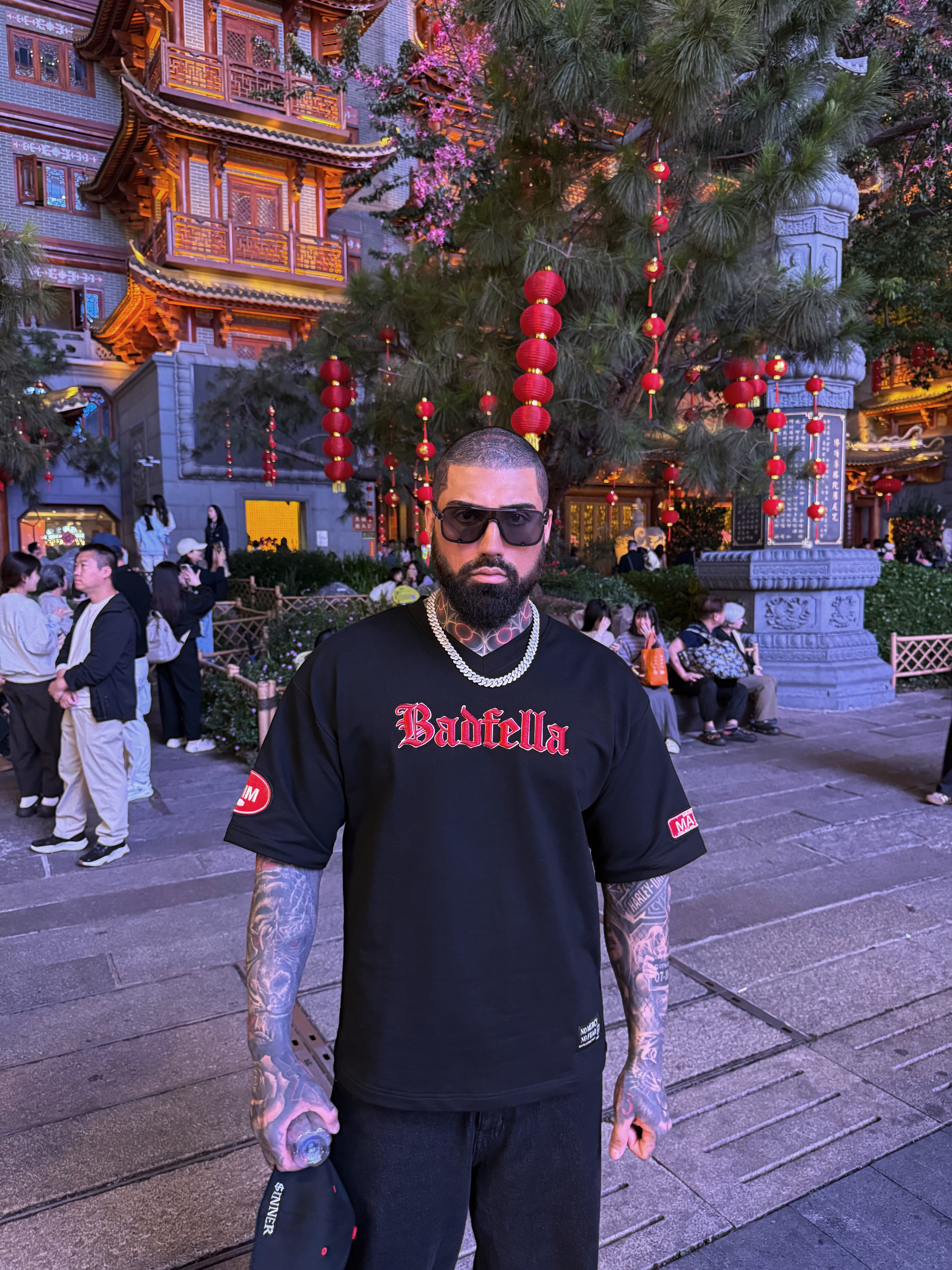 Man wearing a black 'Badfella' shirt in an outdoor setting with traditional architecture and decorations.