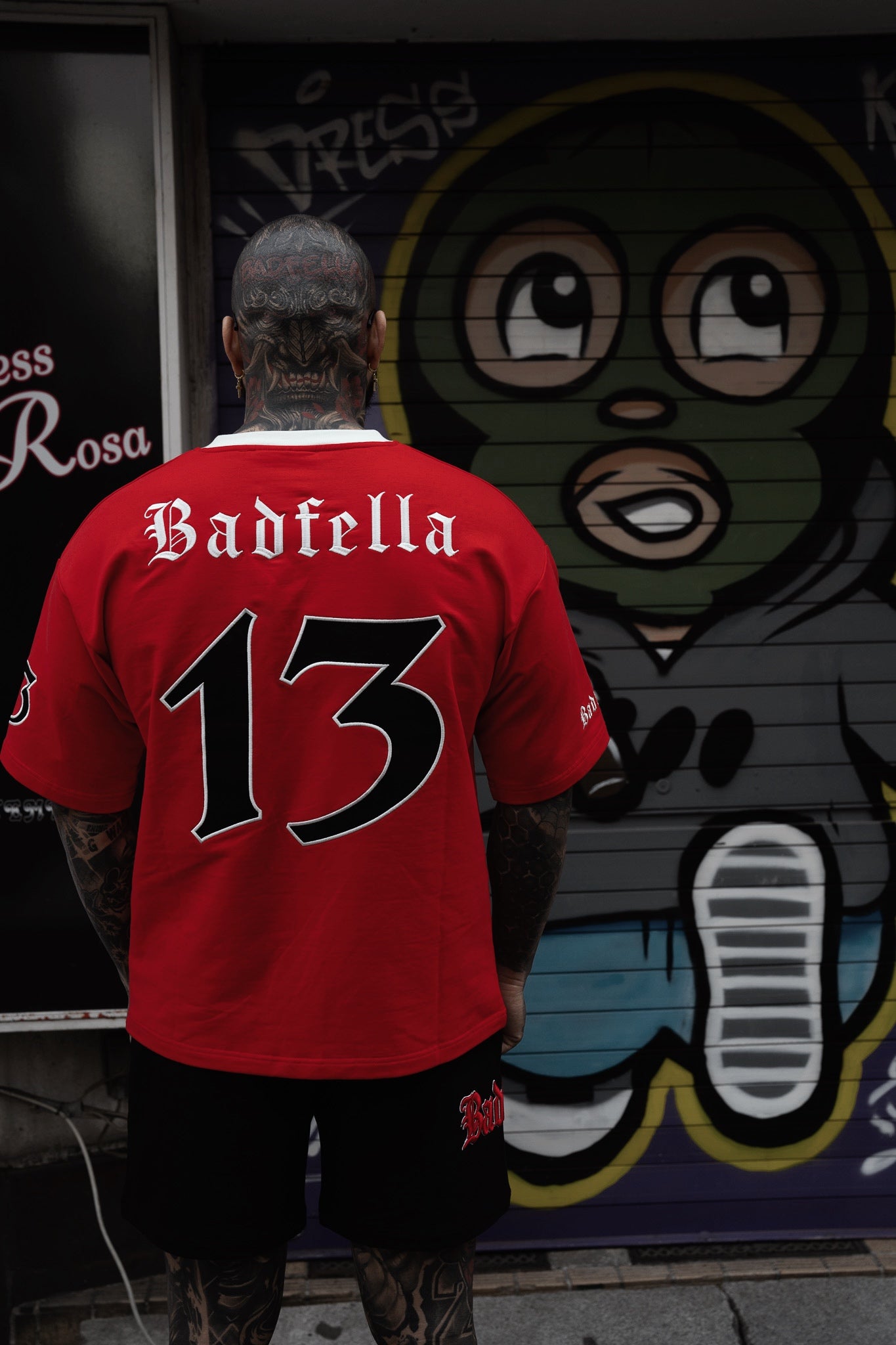 Person wearing a red sports jersey with 'Badfella' and number '13' in front of a mural.