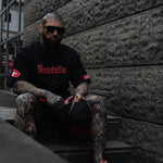 Man with tattoos wearing a black tee with red text, sitting on steps against a textured wall.