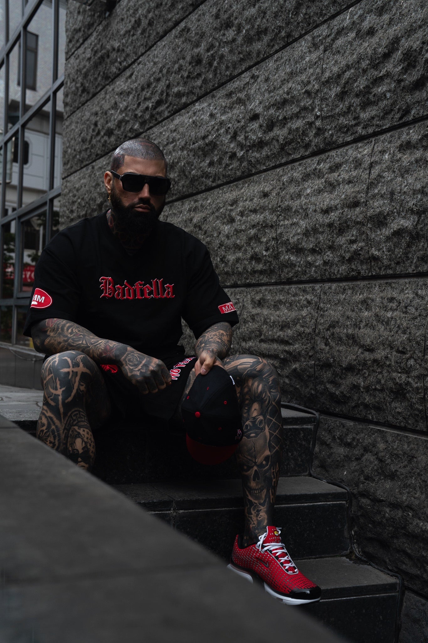 Man with tattoos wearing a black tee with red text, sitting on steps against a textured wall.