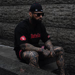 Man with tattoos wearing a black hoodie and cap, sitting against a stone wall.