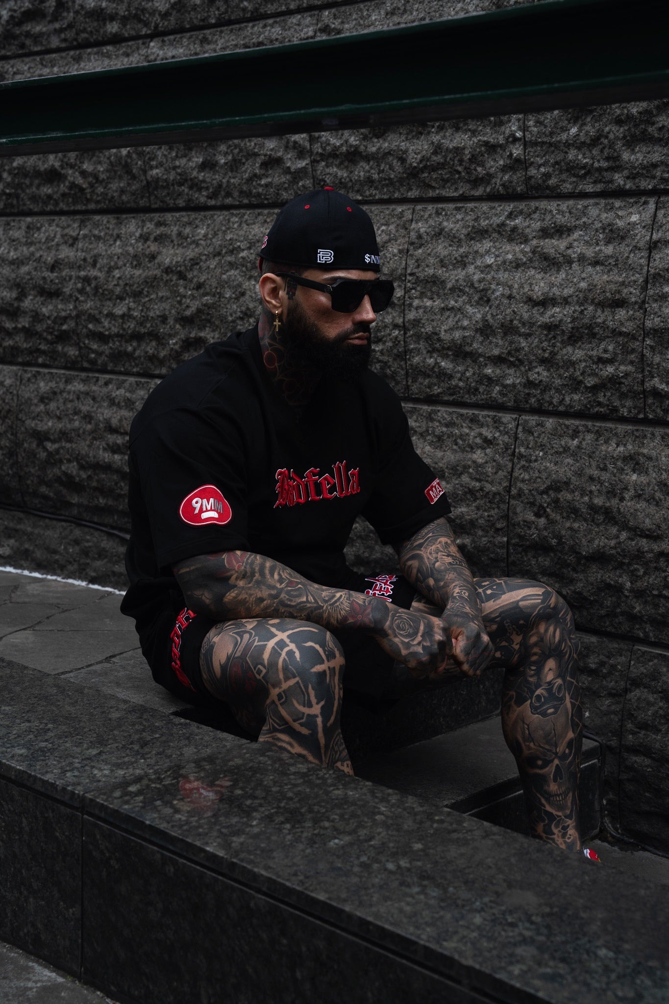 Man with tattoos wearing a black hoodie and cap, sitting against a stone wall.