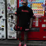 Person wearing a black t-shirt with red text and black shorts with a skull design, standing in front of a colorful vending machine.