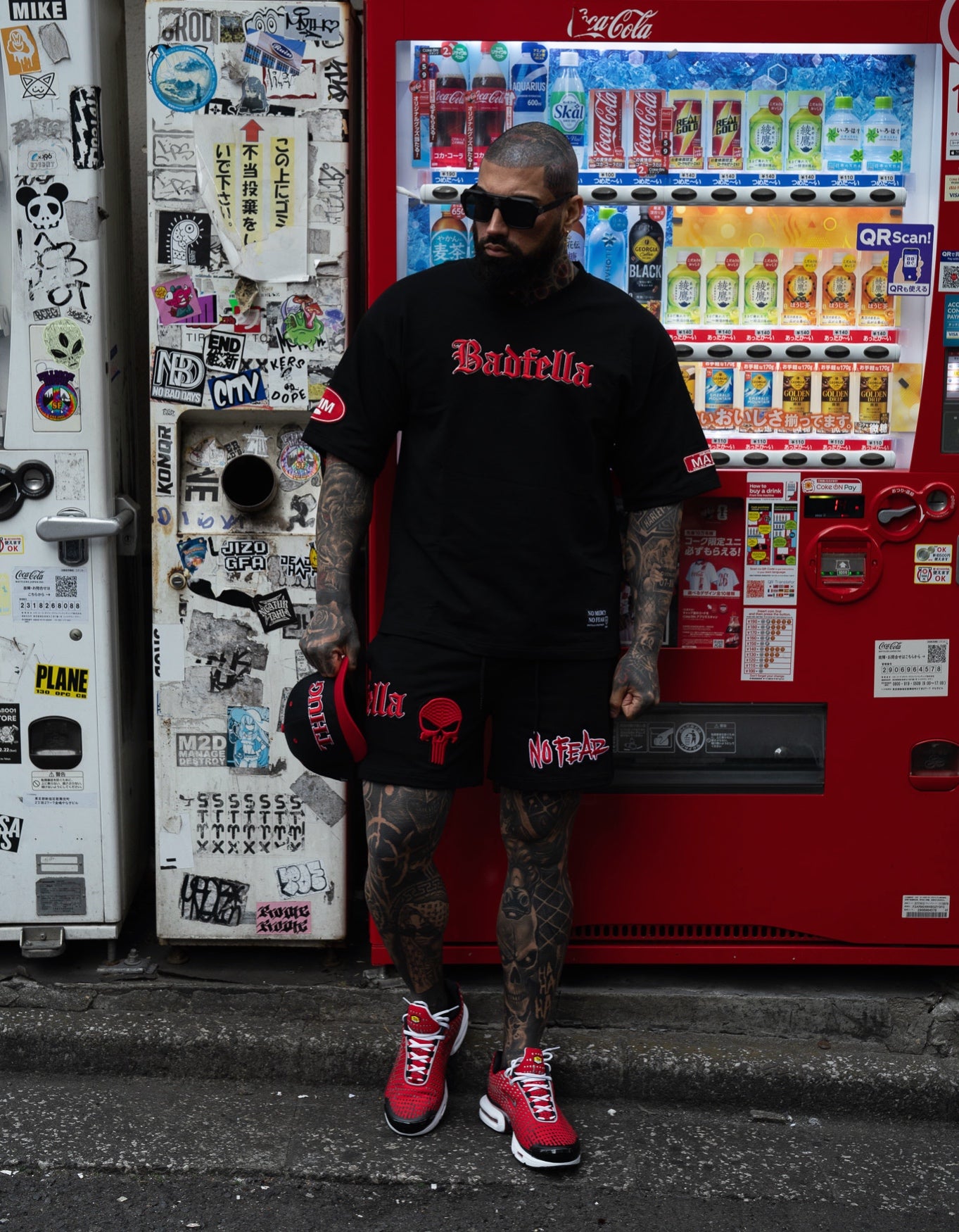 Person wearing a black t-shirt with red text and black shorts with a skull design, standing in front of a colorful vending machine.