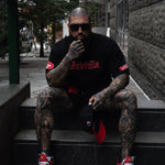 Man with tattoos wearing a black outfit with red accents and red sneakers sitting on steps.