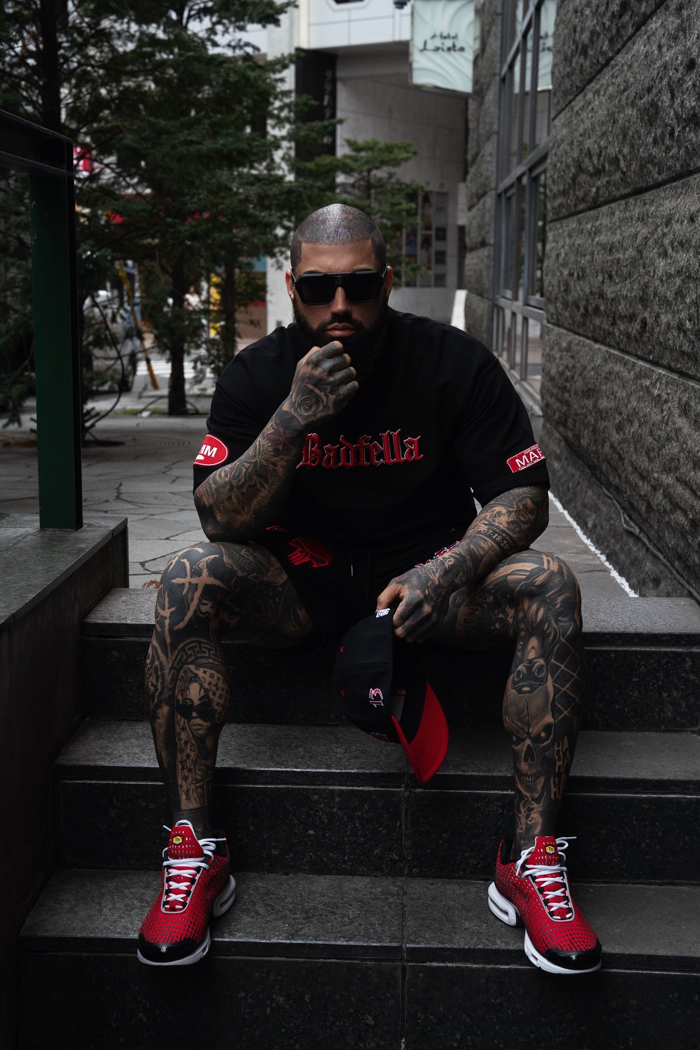 Man with tattoos wearing a black outfit with red accents and red sneakers sitting on steps.